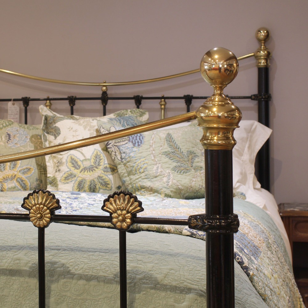 An antique Victorian black painted iron king size bedstead featuring bowed brass top rails, brass ball finials, and cast shell rosette embellishments on the footboard spindles. View II