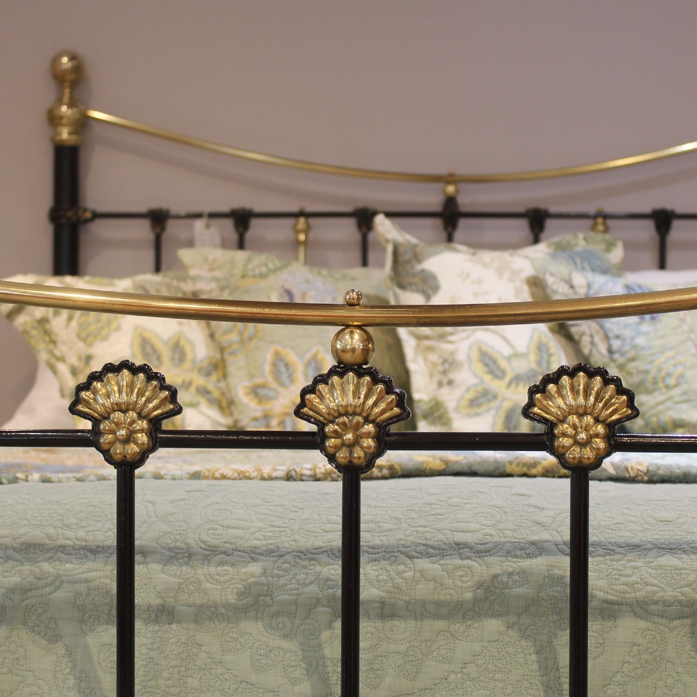 An antique Victorian black painted iron king size bedstead featuring bowed brass top rails, brass ball finials, and cast shell rosette embellishments on the footboard spindles. View III