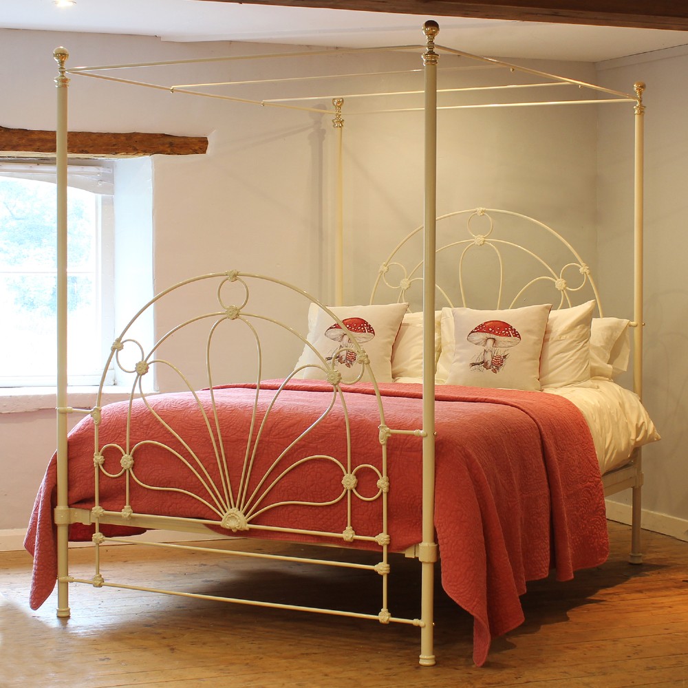 A cream-painted metal four-poster bed frame featuring an arched sunburst motif on the headboard and footboard topped with brass finials.