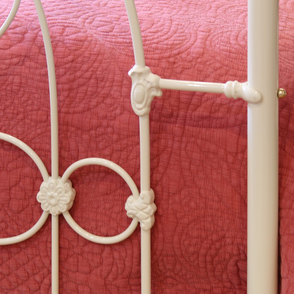 A cream-painted metal four-poster bed frame featuring an arched sunburst motif on the headboard and footboard topped with brass finials. View V