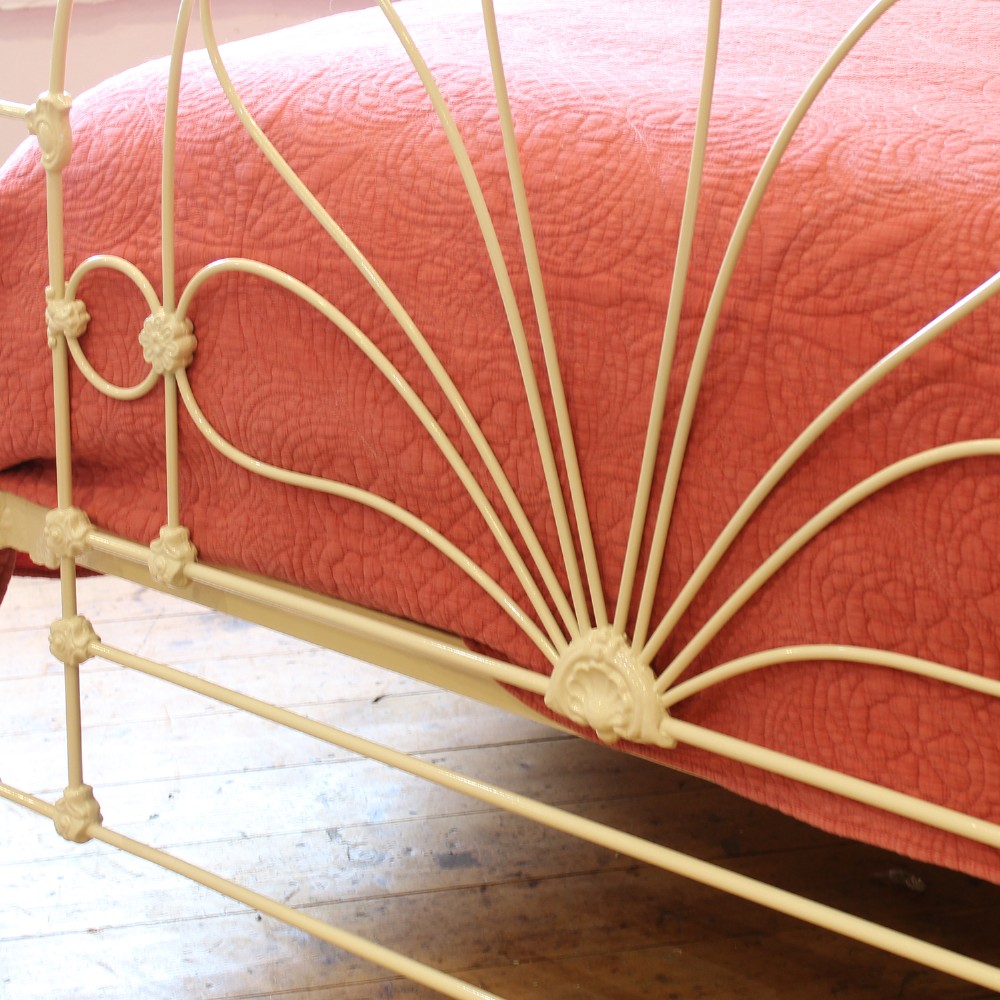 A cream-painted metal four-poster bed frame featuring an arched sunburst motif on the headboard and footboard topped with brass finials. View VI