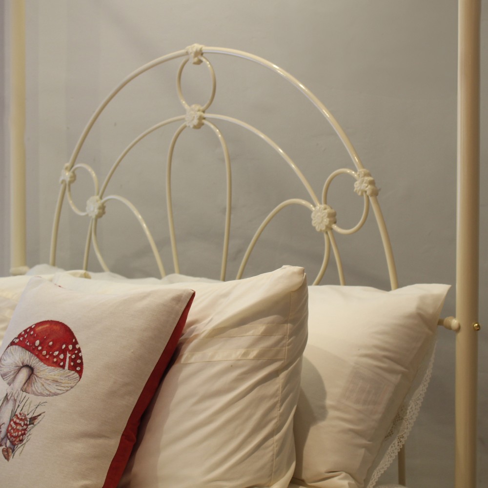 A cream-painted metal four-poster bed frame featuring an arched sunburst motif on the headboard and footboard topped with brass finials. View VII