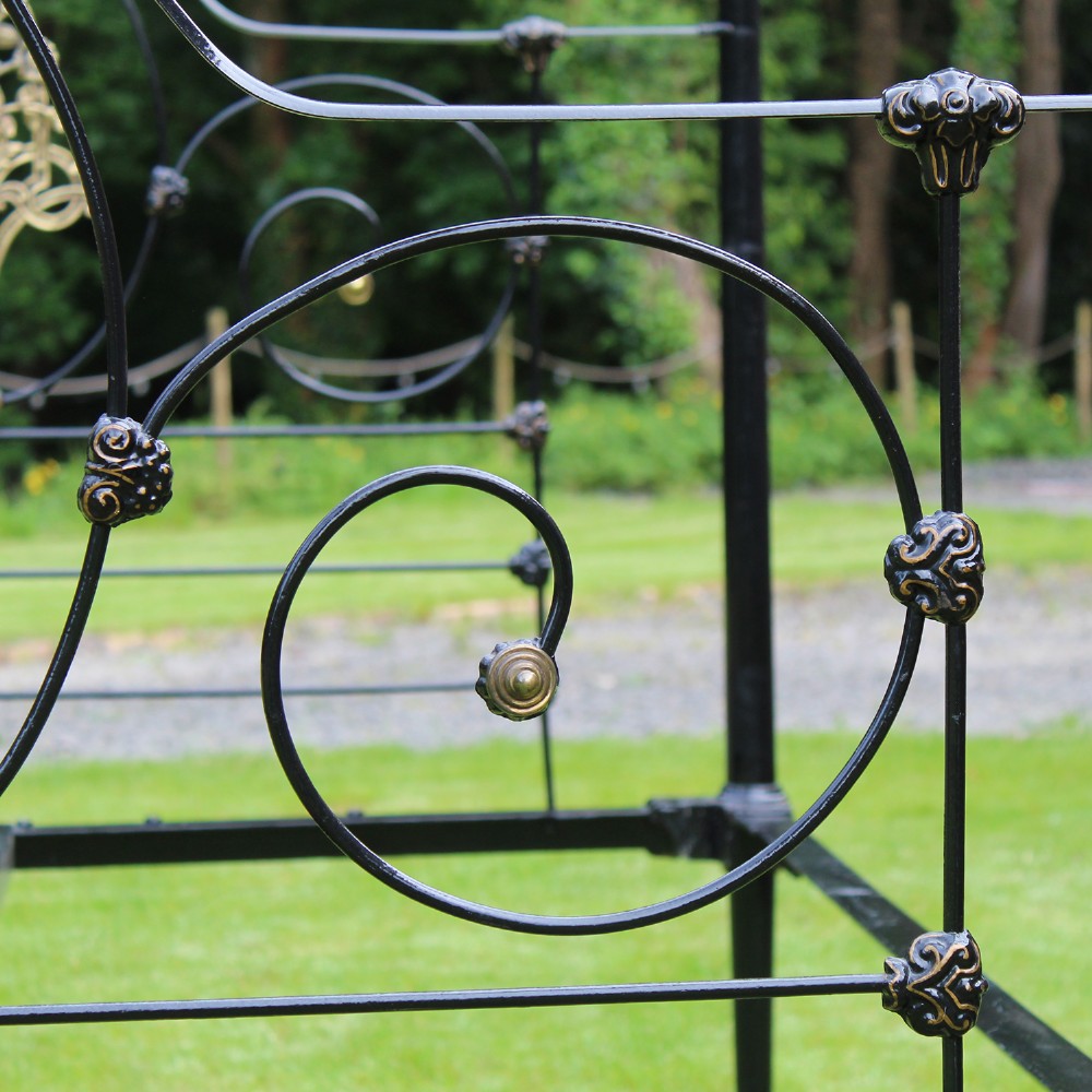 An antique black iron four-poster canopy bed frame featuring decorative scrollwork, central brass urn medallions, and brass finials. View V
