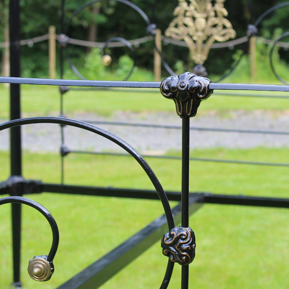 An antique black iron four-poster canopy bed frame featuring decorative scrollwork, central brass urn medallions, and brass finials. View VI