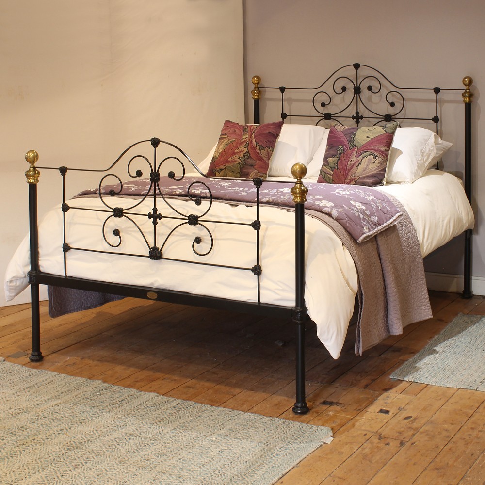 A black wrought iron king-size bed frame featuring decorative scrollwork on the headboard and footboard, topped with brass corner finials.