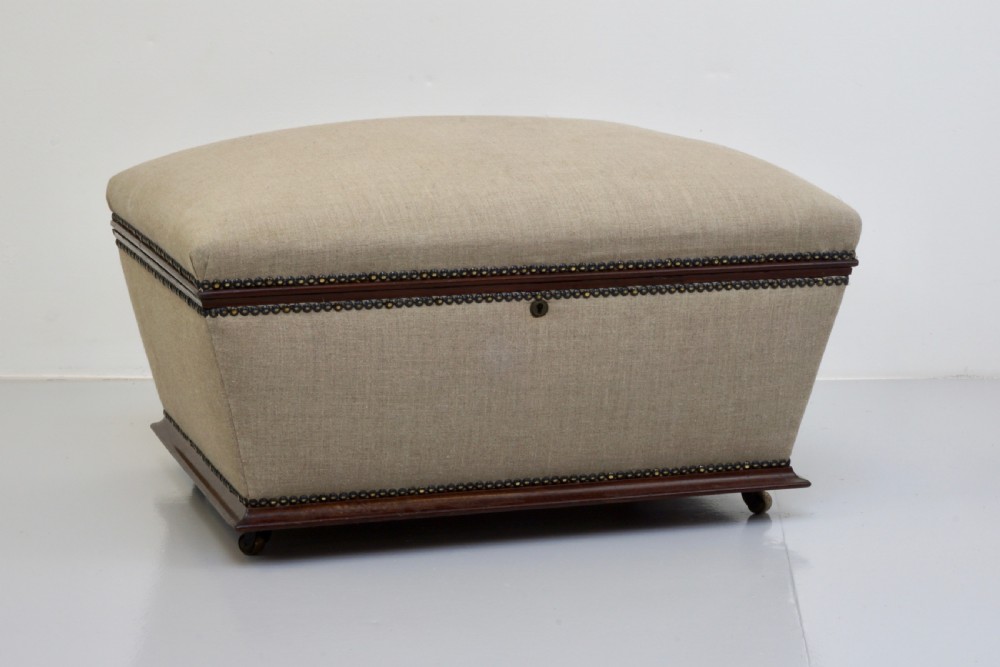 A neutral linen-upholstered antique box ottoman featuring dark wood base trim, brass stud detailing, a front keyhole, and small castors.