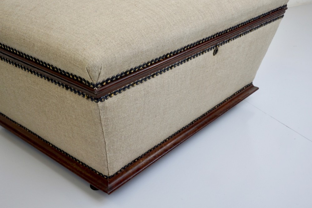 A neutral linen-upholstered antique box ottoman featuring dark wood base trim, brass stud detailing, a front keyhole, and small castors. View II