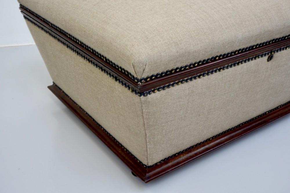 A neutral linen-upholstered antique box ottoman featuring dark wood base trim, brass stud detailing, a front keyhole, and small castors. View III