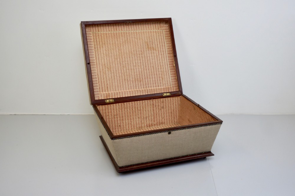 A neutral linen-upholstered antique box ottoman featuring dark wood base trim, brass stud detailing, a front keyhole, and small castors. View IV