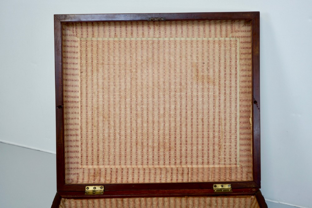 A neutral linen-upholstered antique box ottoman featuring dark wood base trim, brass stud detailing, a front keyhole, and small castors. View V