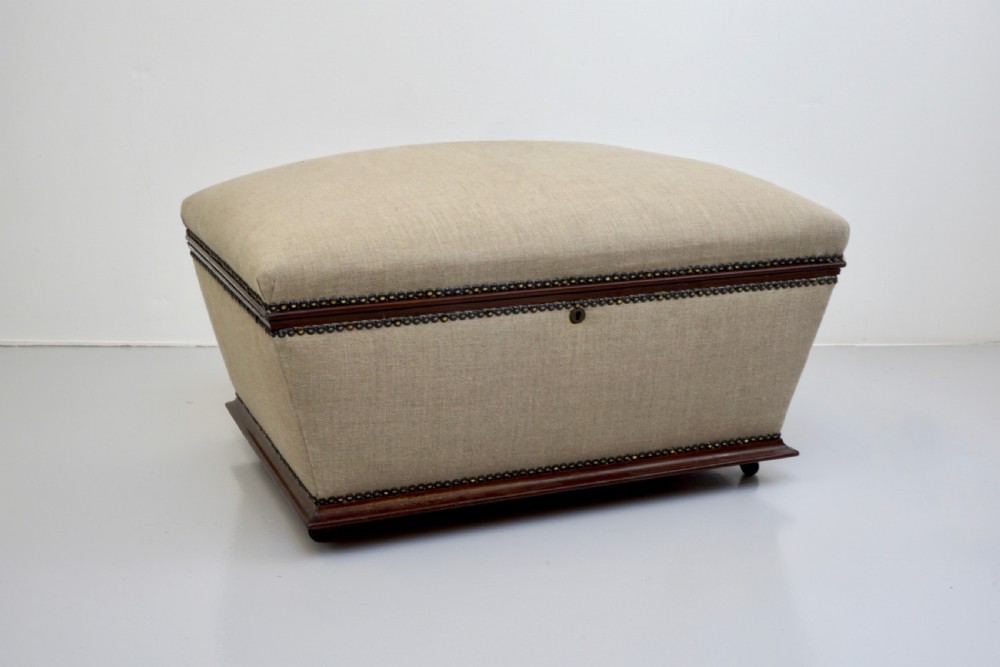 A neutral linen-upholstered antique box ottoman featuring dark wood base trim, brass stud detailing, a front keyhole, and small castors. View VI