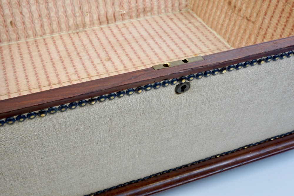 A neutral linen-upholstered antique box ottoman featuring dark wood base trim, brass stud detailing, a front keyhole, and small castors. View VII