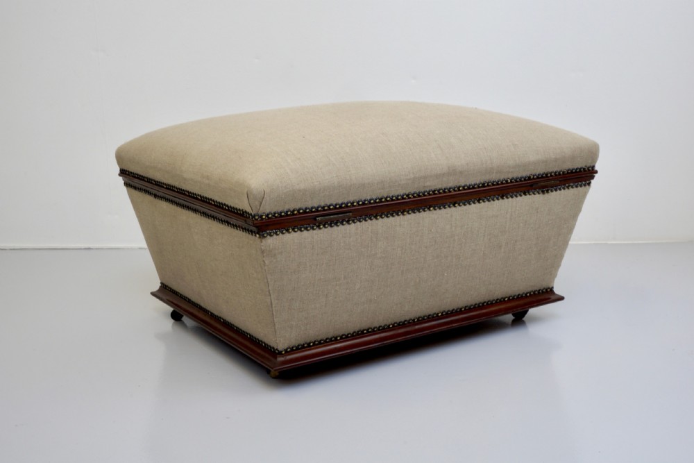 A neutral linen-upholstered antique box ottoman featuring dark wood base trim, brass stud detailing, a front keyhole, and small castors. View IX