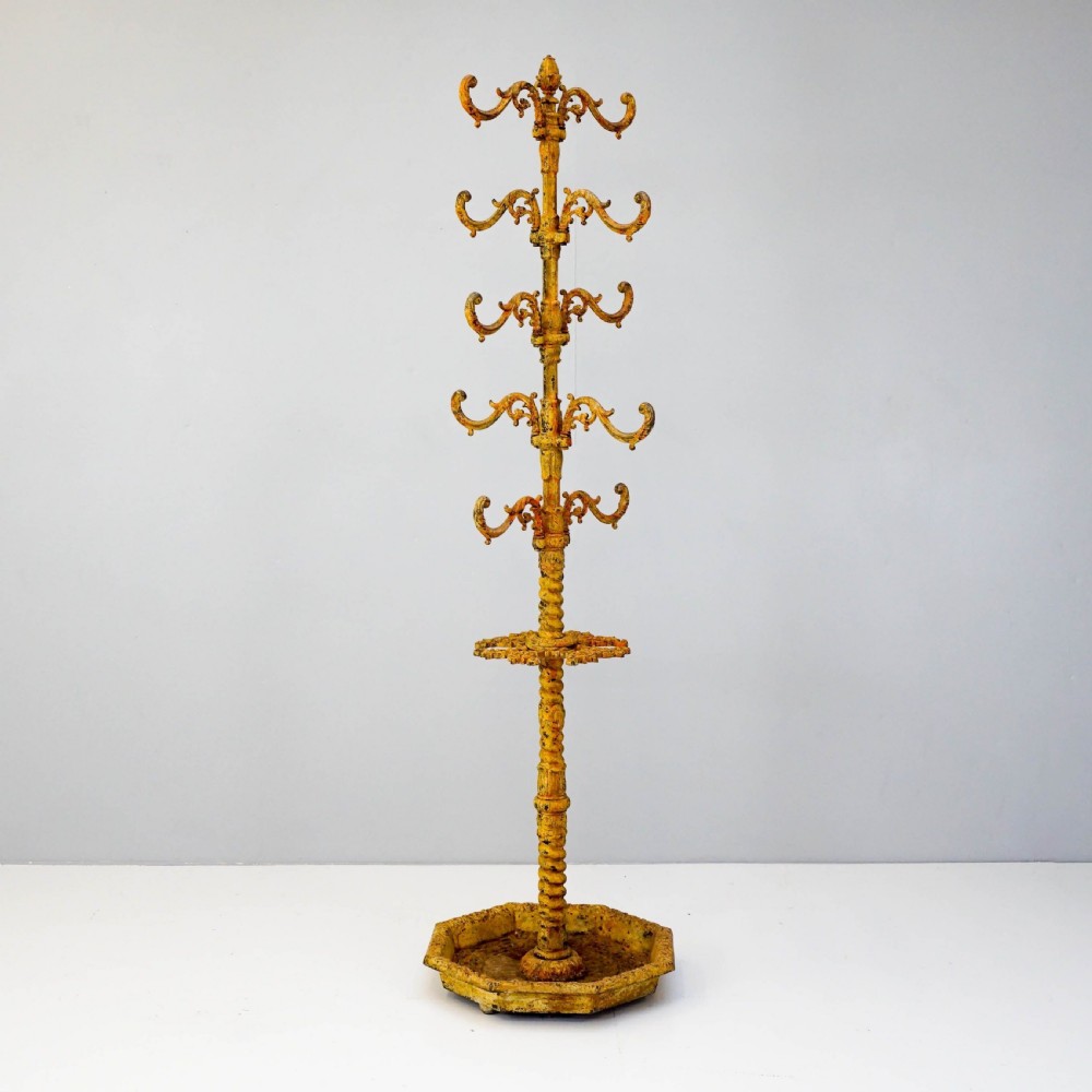 A tall, ornate yellow-painted cast iron coat stand featuring multiple tiers of scroll hooks, a twisted central column, and a hexagonal drip tray base.