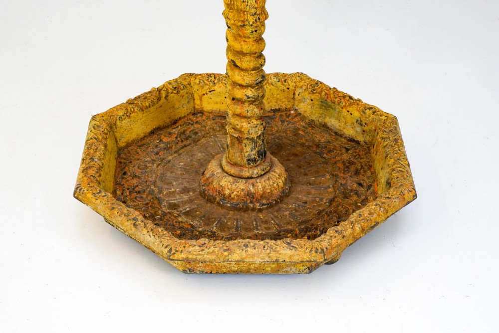 A tall, ornate yellow-painted cast iron coat stand featuring multiple tiers of scroll hooks, a twisted central column, and a hexagonal drip tray base. View VII
