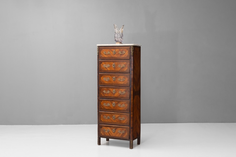 A tall, narrow French semainier chest of drawers featuring geometric wood parquetry, seven drawers, gilt bronze hardware, and a white marble top.