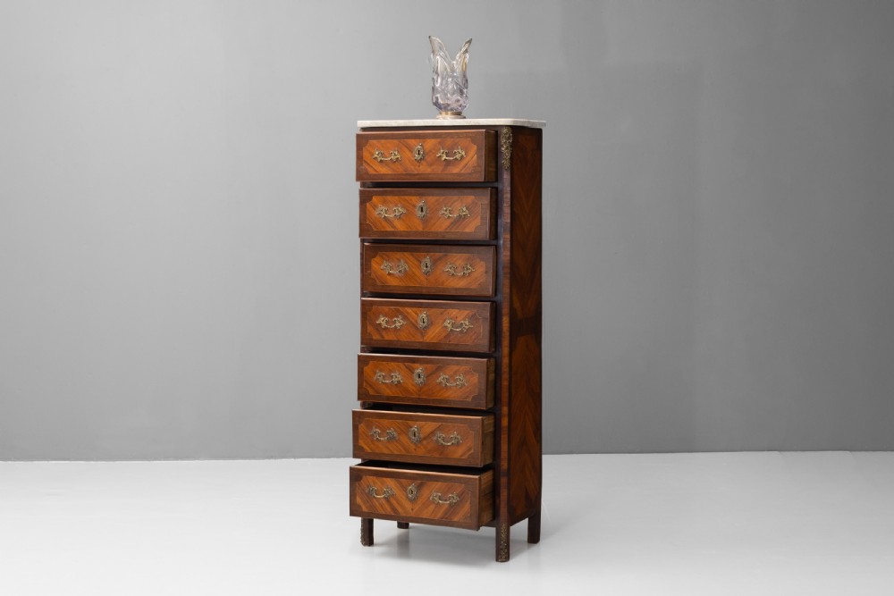 A tall, narrow French semainier chest of drawers featuring geometric wood parquetry, seven drawers, gilt bronze hardware, and a white marble top. View II