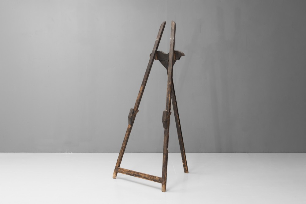 A tall, dark patinated pine A-frame studio easel featuring adjustable rest pegs and a carved decorative upper crossbar.
