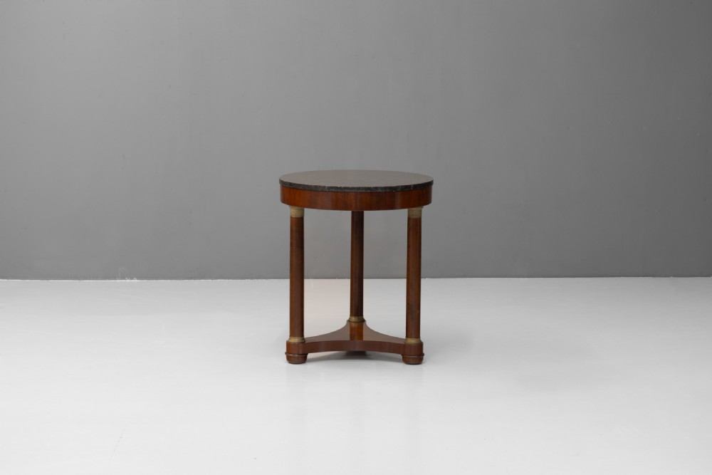 A French Empire style circular mahogany gueridon table featuring a dark marble top supported by three cylindrical columns on a tri-form base.
