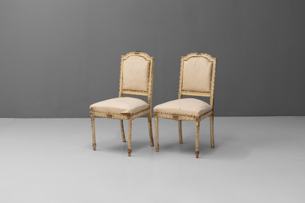 A pair of French Louis XVI style side chairs featuring distressed cream painted frames, tapered fluted legs, and neutral fabric upholstery.