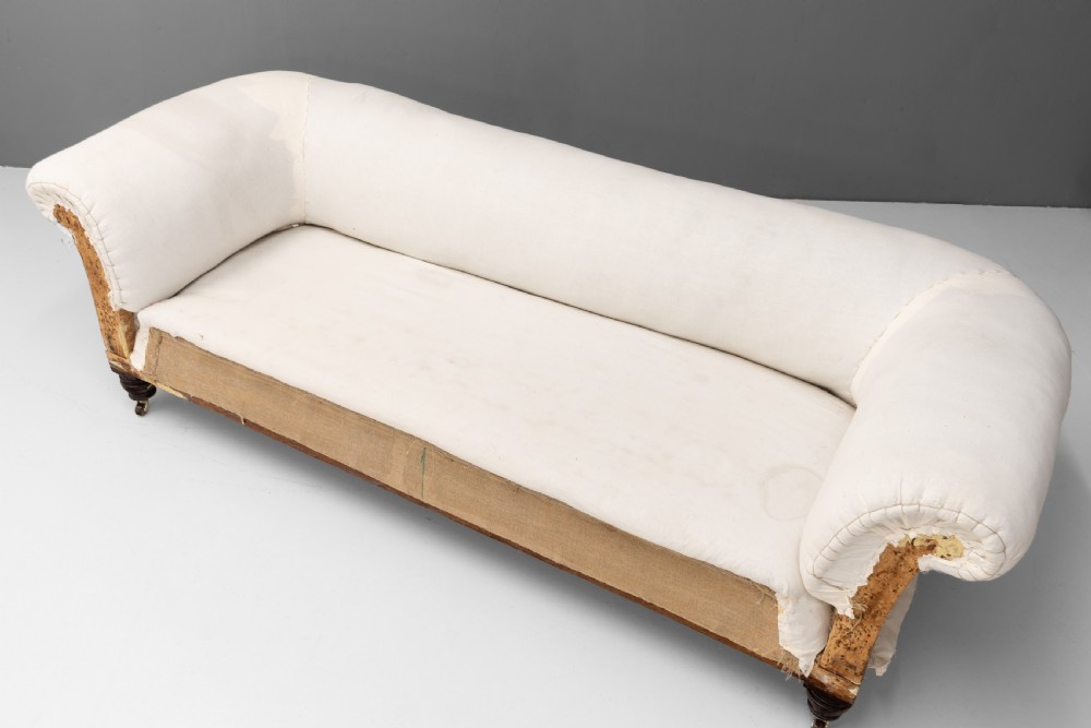 An antique Victorian Chesterfield settee upholstered in off-white calico lining, featuring deep rolled arms, exposed hessian lower upholstery, and turned mahogany legs with brass castors. View IV