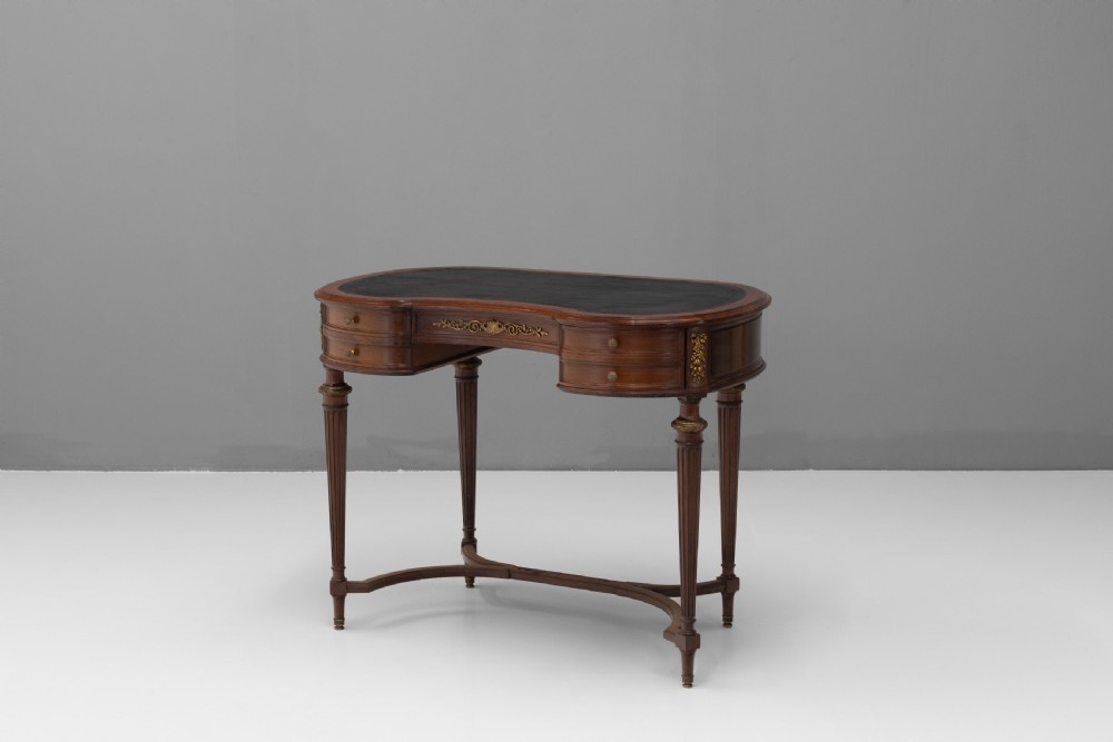 A mahogany kidney-shaped writing desk with a dark leather inset top, gilt-bronze mounts, four side drawers, and fluted legs joined by a curved stretcher.
