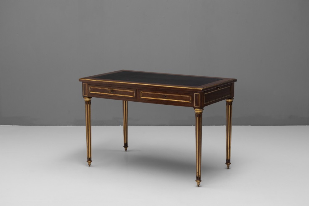 A French Louis XVI style mahogany bureau plat featuring a dark leather writing insert, gilt-bronze trim, frieze drawers, and fluted tapered legs.