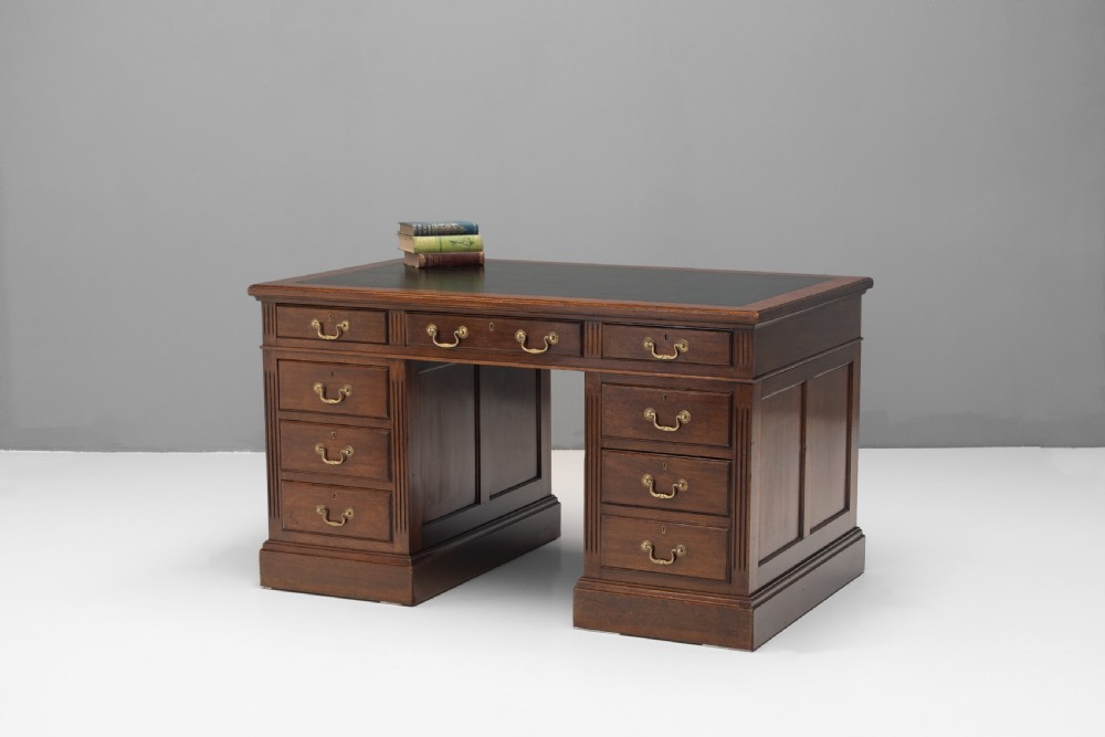 An antique mahogany pedestal desk featuring a dark leather top, fluted side columns, nine drawers with brass handles, and a central kneehole.