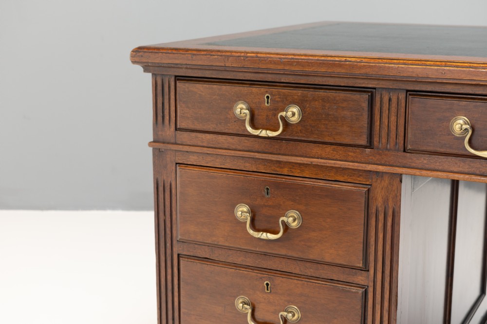 An antique mahogany pedestal desk featuring a dark leather top, fluted side columns, nine drawers with brass handles, and a central kneehole. View IV