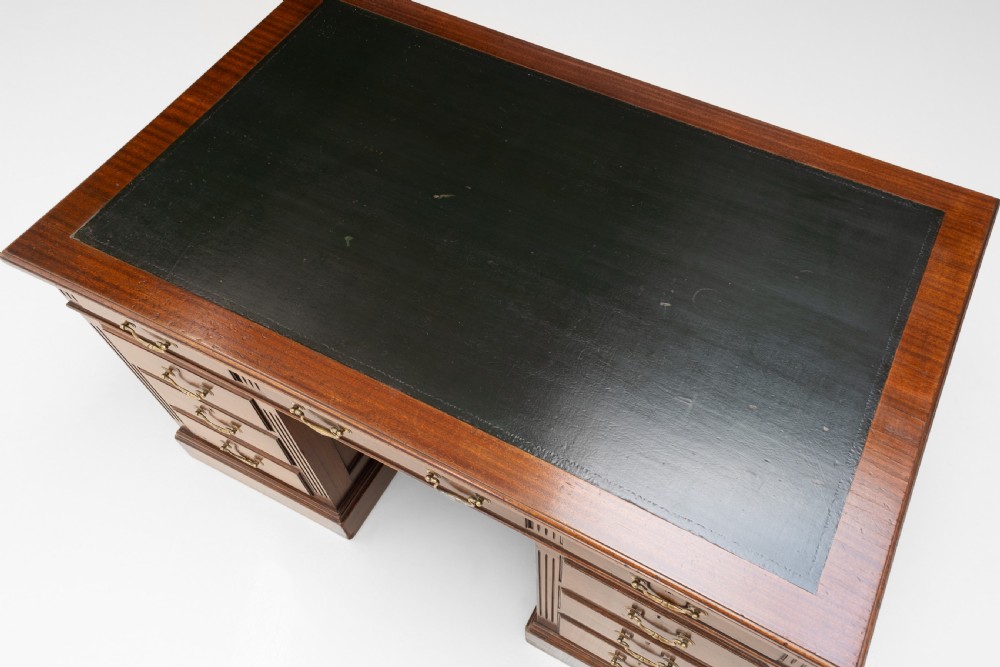 An antique mahogany pedestal desk featuring a dark leather top, fluted side columns, nine drawers with brass handles, and a central kneehole. View V