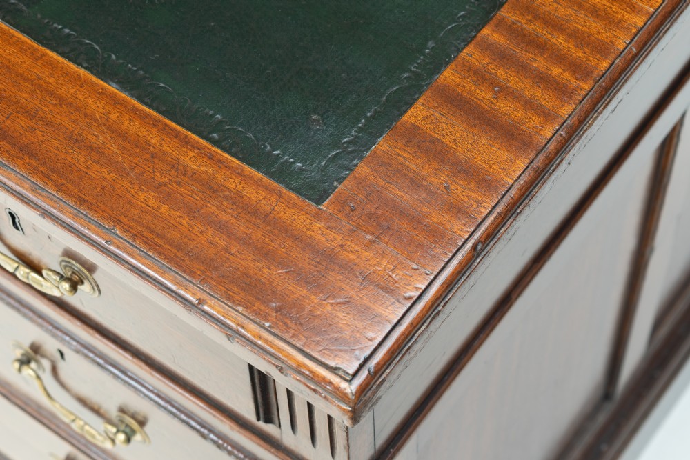 An antique mahogany pedestal desk featuring a dark leather top, fluted side columns, nine drawers with brass handles, and a central kneehole. View VI