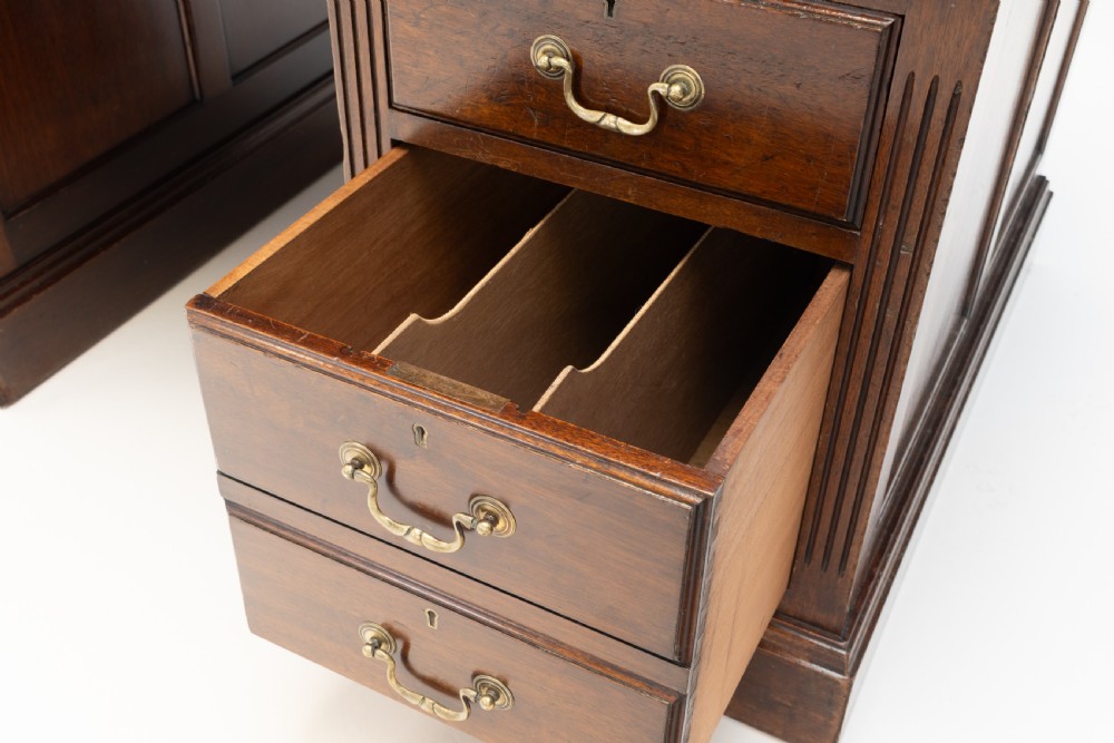An antique mahogany pedestal desk featuring a dark leather top, fluted side columns, nine drawers with brass handles, and a central kneehole. View VII