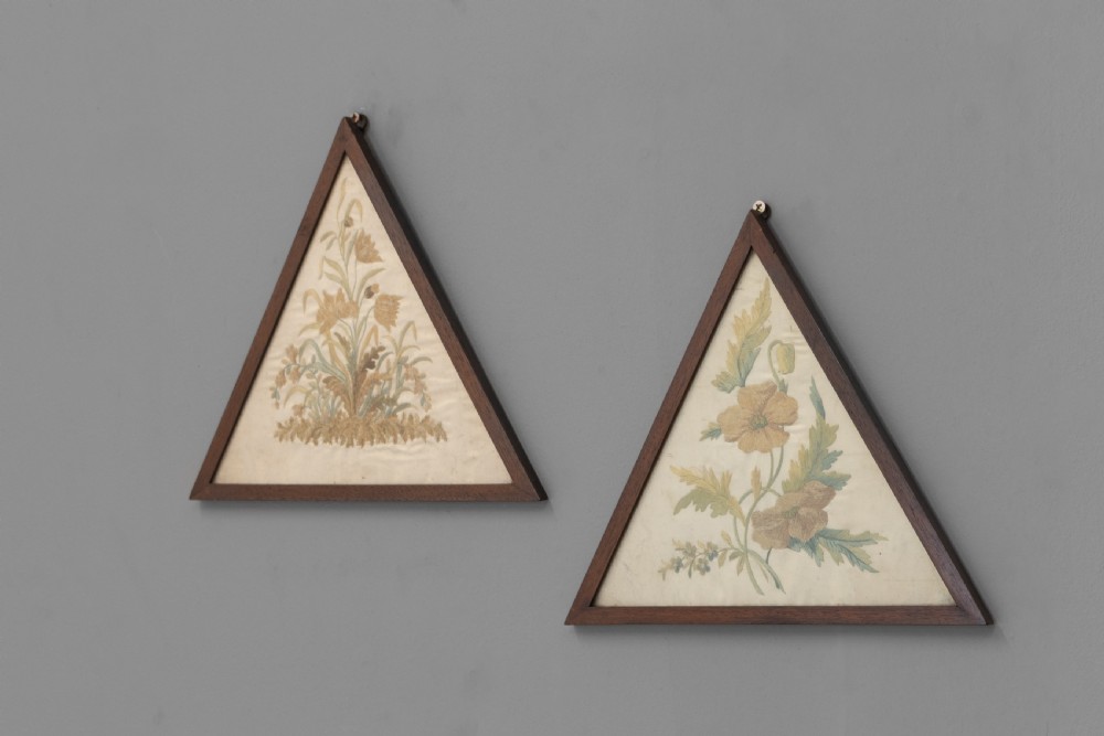 A pair of triangular wooden frames displaying hand-stitched botanical embroidery featuring floral motifs on a cream fabric ground.