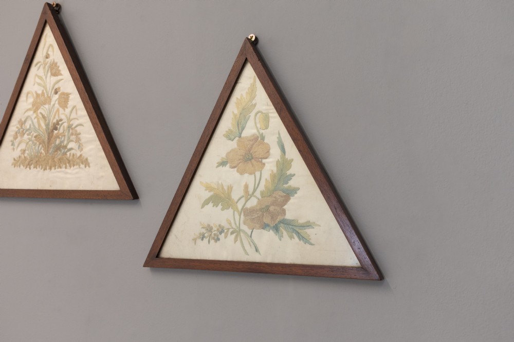 A pair of triangular wooden frames displaying hand-stitched botanical embroidery featuring floral motifs on a cream fabric ground. View II