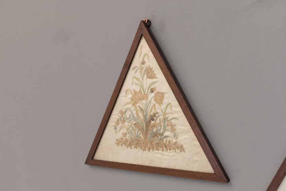 A pair of triangular wooden frames displaying hand-stitched botanical embroidery featuring floral motifs on a cream fabric ground. View III