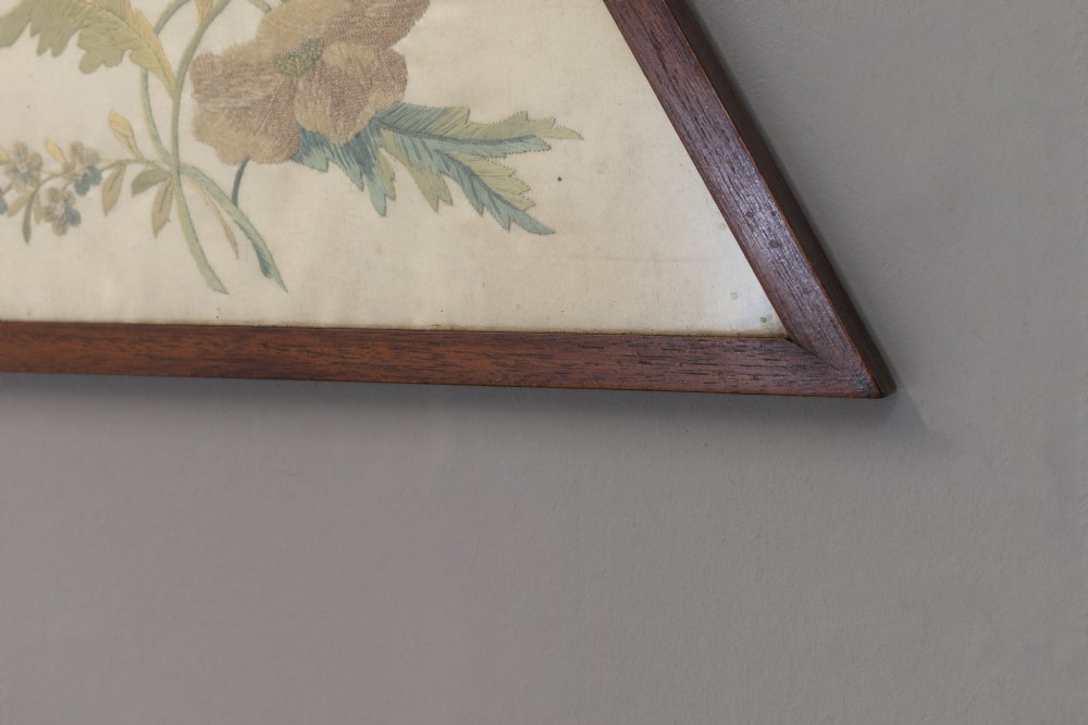 A pair of triangular wooden frames displaying hand-stitched botanical embroidery featuring floral motifs on a cream fabric ground. View V