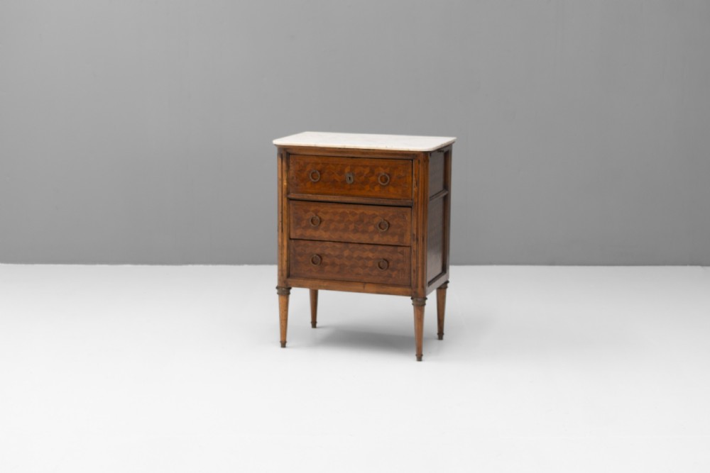 A Neoclassical style three-drawer wooden chest featuring geometric parquetry inlay, brass ring handles, tapered legs, and a white marble top surface.