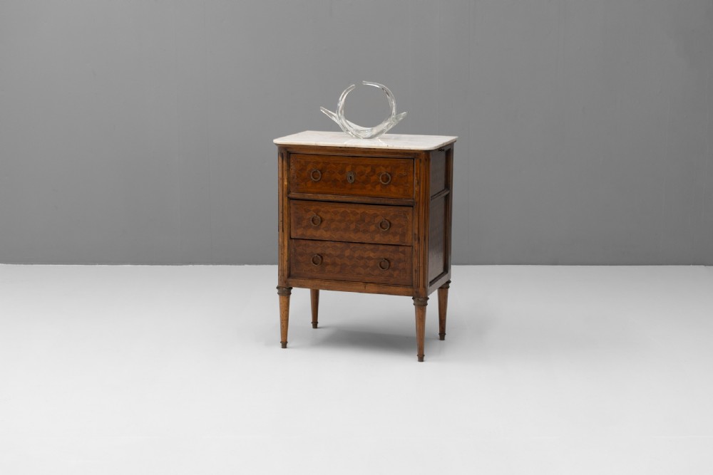 A Neoclassical style three-drawer wooden chest featuring geometric parquetry inlay, brass ring handles, tapered legs, and a white marble top surface. View IV