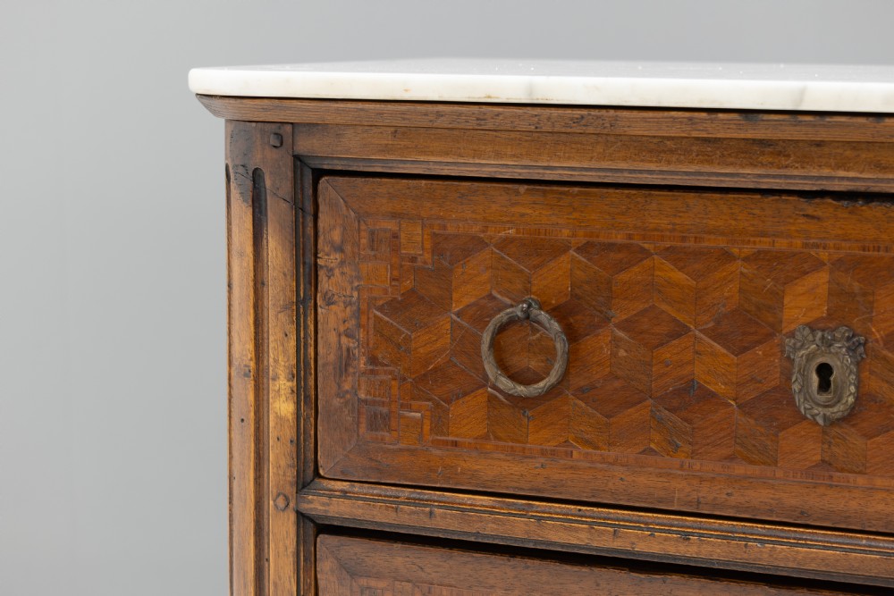 A Neoclassical style three-drawer wooden chest featuring geometric parquetry inlay, brass ring handles, tapered legs, and a white marble top surface. View VI
