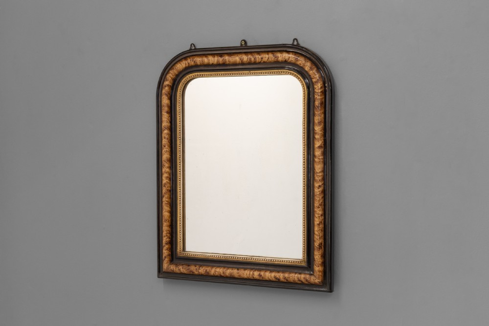 A small Louis Philippe wall mirror featuring rounded upper corners, an ebonised outer profile, figured wood veneer, and a beaded gilt inner border.