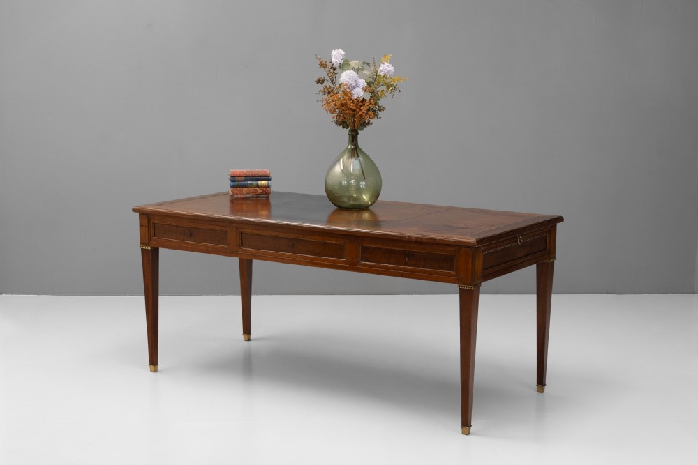 A rectangular mahogany writing table with an inset brown leather top, three frieze drawers, and four square tapered legs ending in brass caps.