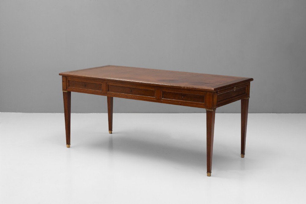 A rectangular mahogany writing table with an inset brown leather top, three frieze drawers, and four square tapered legs ending in brass caps. View II