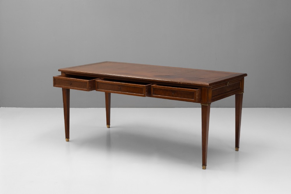A rectangular mahogany writing table with an inset brown leather top, three frieze drawers, and four square tapered legs ending in brass caps. View III
