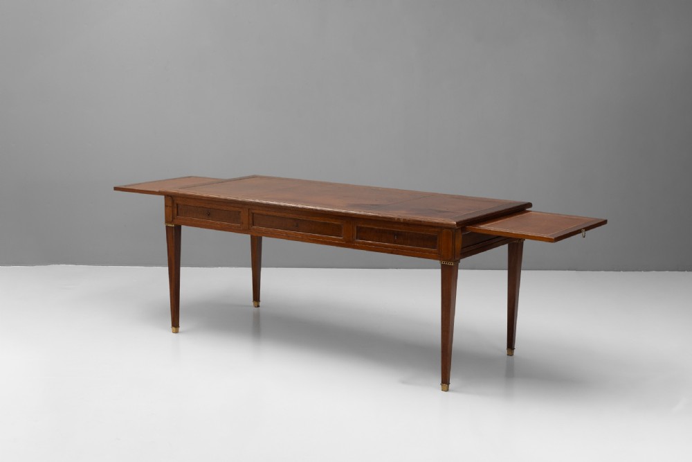 A rectangular mahogany writing table with an inset brown leather top, three frieze drawers, and four square tapered legs ending in brass caps. View IV