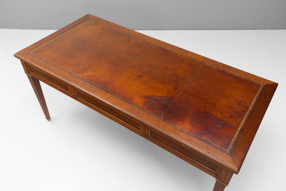 A rectangular mahogany writing table with an inset brown leather top, three frieze drawers, and four square tapered legs ending in brass caps. View V