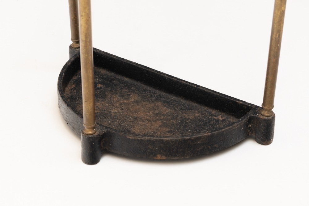 A semi-circular brass stick stand featuring tubular uprights topped with turned finials and supported by a heavy black cast iron drip tray base. View V