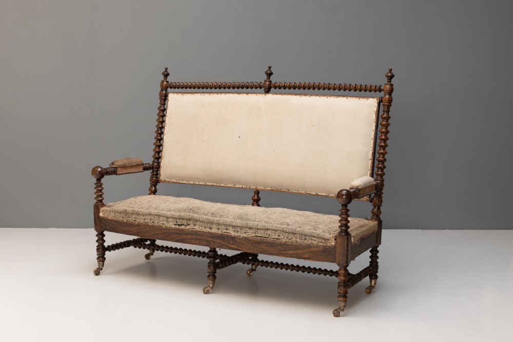 A 19th-century French bobbin-turned oak settee featuring a high upholstered back panel, spool-turned wooden framing, and brass castors.