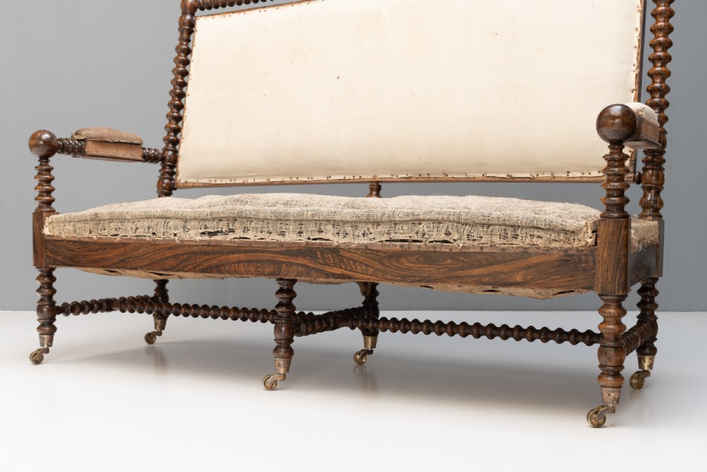 A 19th-century French bobbin-turned oak settee featuring a high upholstered back panel, spool-turned wooden framing, and brass castors. View VI