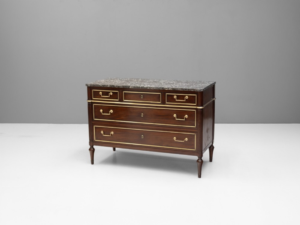 A dark mahogany French Louis XVI style chest of drawers featuring a grey marble top, brass linear moulding, brass swing handles, and turned tapered legs.