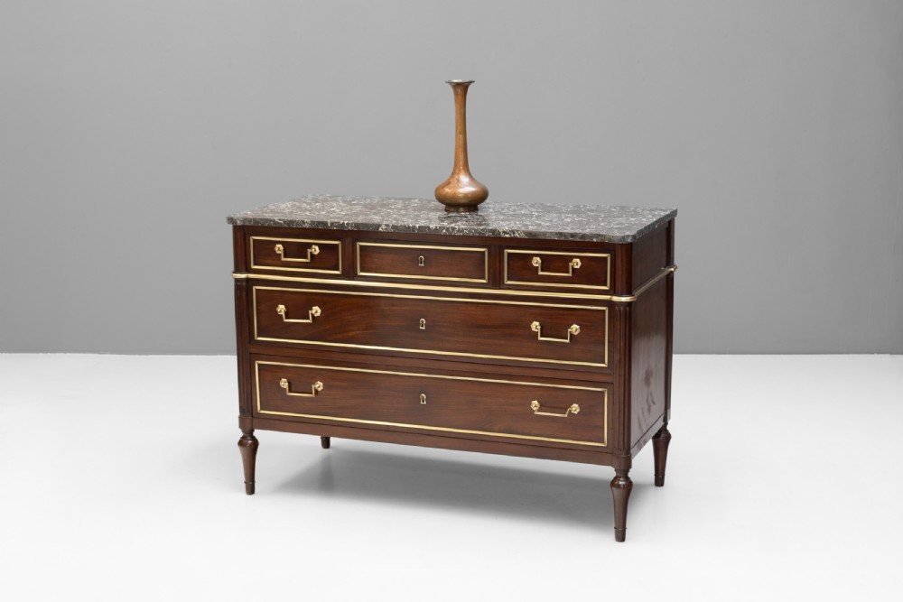 A dark mahogany French Louis XVI style chest of drawers featuring a grey marble top, brass linear moulding, brass swing handles, and turned tapered legs. View II